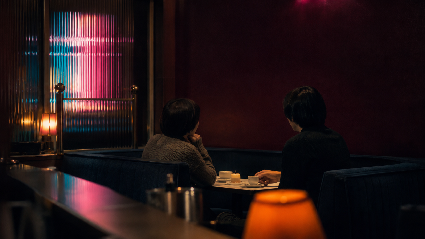 夜のボックス席で向かい合う二人の後ろ姿。卓上のキャンドルと珈琲、奥の窓にはネオンの色が縞になって映っている。