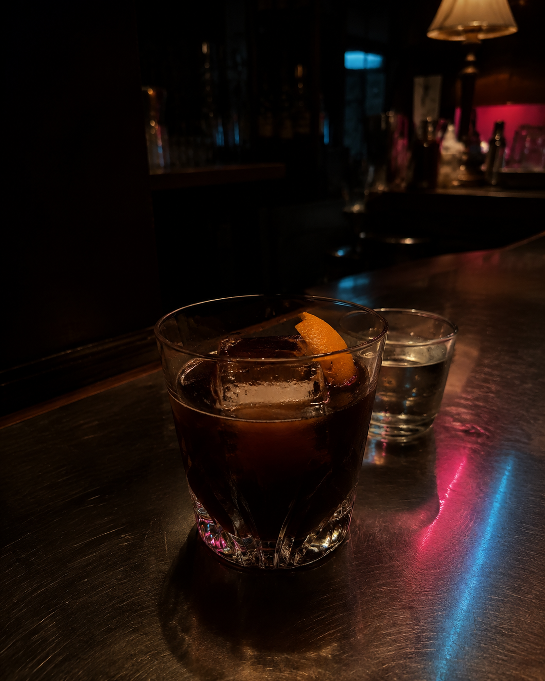 暗いカウンターに置かれたロックグラス。大きな氷とオレンジピールが入った珈琲とウイスキーの一杯、奥にチェイサーの水。